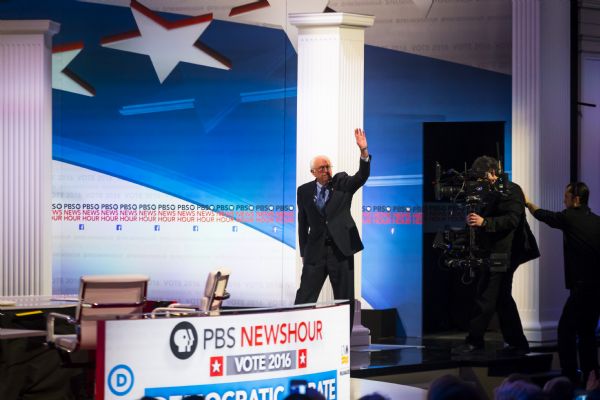 The U.S. Senator from Vermont, Bernie Sanders, waving to the crowd as he is walking on stage for the Democratic presidential debate. A camera operator is standing behind him.