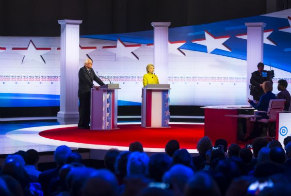 Hillary Clinton and Bernie Sanders standing at podiums and answering questions posed by Gwen Ifill and Judy Woodruff, the moderators of the Democratic presidential debate. A camera operator is filming in the background and a crowd is watching from seats below the stage.