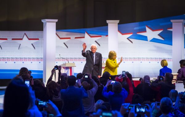During a commercial break from the television broadcast Democratic presidential debate hosted by PBS News Hour, candidates Bernie Sanders and Hillary Clinton are waving to supporters. People near the stage are standing to take pictures of the two candidates. The moderators, Gwen Ifill and Judy Woodruff, are sitting at their desk on the right.