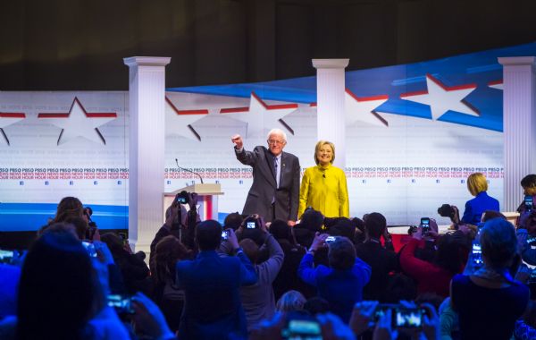 During a commercial break from the television broadcast of the Democratic presidential debate hosted by PBS News Hour, candidates Bernie Sanders and Hillary Clinton are standing and smiling at supporters. The two moderators, Gwen Ifill and Judy Woodruff, are sitting at their desk on the right.