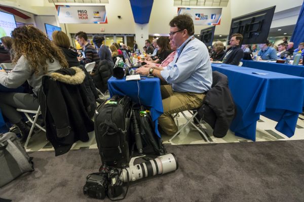 Side view of journalists and reporters sitting at long tables watching the debate and working on computers or taking notes. In the foreground, Mark Hoffman, a photojournalist for the "Milwaukee Journal Sentinel," is sitting at his computer with his professional camera equipment on the floor next to him.