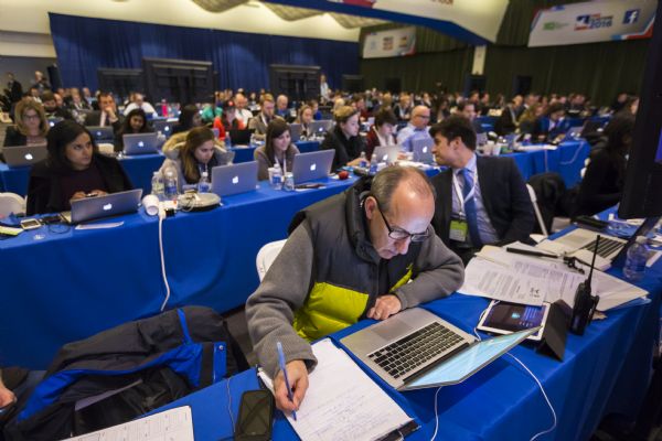View of journalists sitting behind long tables working in the media filing center during the debate. A man sitting in the foreground is looking down at his computer and taking notes on a legal pad. The man next to him is leaning back to talk to a woman working on her computer behind him.