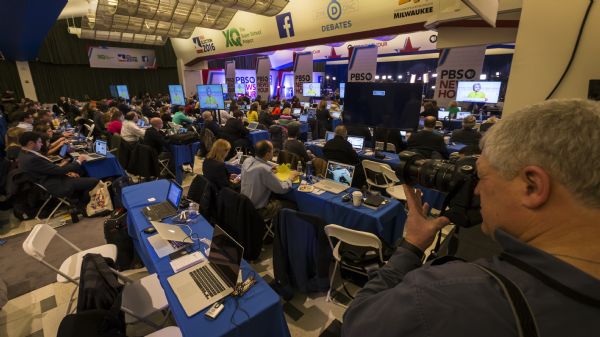 Slightly elevated view of the journalists and reporters in the media room at the Democratic presidential debate. In the right foreground is Associated Press freelance photojournalist Tom Lynn taking a photograph. The journalists are sitting at long tables while working on computers or taking notes while watching Hillary Clinton on the screens broadcasting the debates.