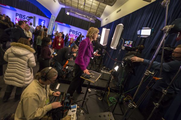 U.S. Senator Tammy Baldwin of Wisconsin, a Hillary Clinton supporter, is being interviewed in the spin room after the Democratic presidential debate. She is standing on a small stage crowded with equipment, journalists, technicians, and a camera operator.