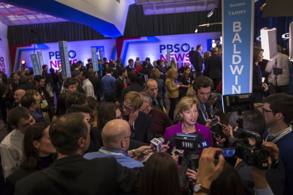 View over crowd of people towards U.S. Senator Tammy Baldwin of Wisconsin, a Hillary Clinton supporter, talking to reporters holding microphones and cameras after the Democratic presidential debate.