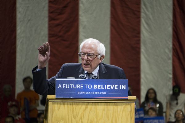 Bernie Sanders standing at a podium during his rally at the Kohl Center. He is waving a hand while addressing the crowd. A group of men and women, mostly young, are standing or sitting behind him. Some of them are holding campaign signs with Sanders' slogan: "A Future to Believe In." There is a large American flag in the background.