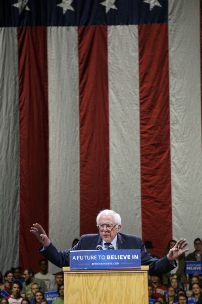 Bernie Sanders standing at a podium, his arms outstretched while addressing the crowed at his rally. A large crowd of people, mostly young, are standing or sitting behind him. Some of them are holding campaign signs bearing his slogan: "A Future to Believe In." There is a large American flag in the background.