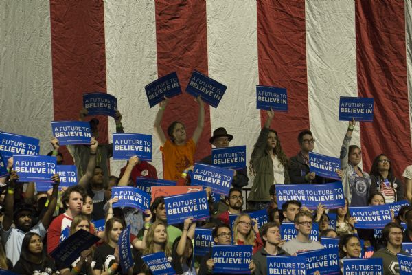 A crowd of people, mostly young, are standing or sitting while holding Bernie Sanders campaign signs. There is a large American flag in the background.