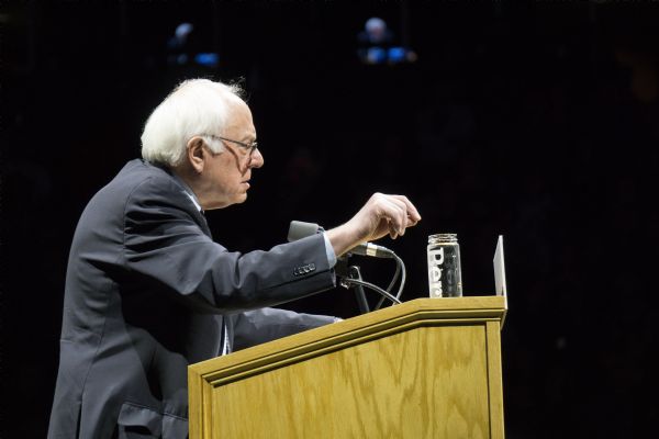 Profile view of Bernie Sanders standing at a podium during his rally at the Kohl Center. He is gesturing with one hand while addressing the crowd. On the podium is an open bottle of water with his name "Bernie" printed on it.