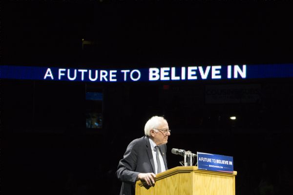 Bernie Sanders standing at the podium at his rally in the Kohl Center. A lighted sign high up in the background is spelling out his campaign slogan: "A Future to Believe In."