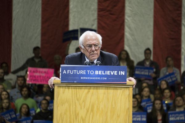 Bernie Sanders standing at the podium during his rally looking over the crowd. Behind him is a group of mostly young men and women, some holding up signs with his campaign slogan: "A Future to Believe In." One person is holding up a bright pink handmade sign reading: "Radical Political Revolution." There is a large American flag in the background.
