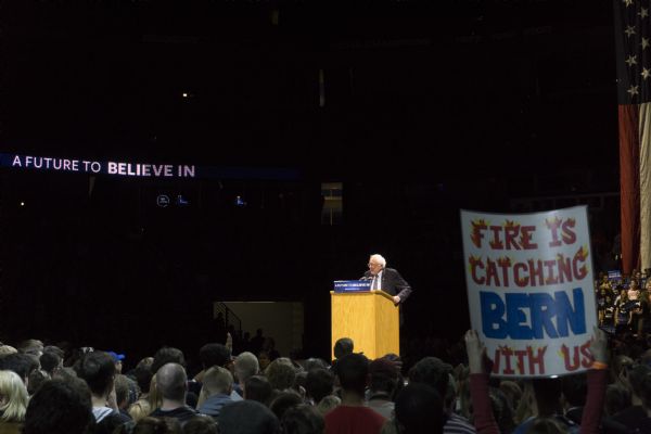 View from the audience at the Bernie Sanders rally held in the Kohl Center. Sanders is standing on a stage behind a podium and is looking out at the crowd. A lighted sign high up in the background is spelling out his campaign slogan: "A Future to Believe In." In the foreground, a woman is holding up a handmade sign reading: "Fire is Catching, Bern with Us."