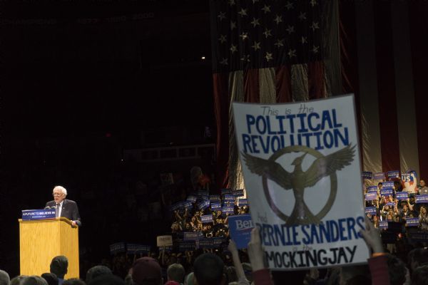 In the foreground someone is holding up a political poster at the Bernie Sanders rally held at the Kohl Center. The sign reads: "This is the Political Revolution and Bernie Sanders is the Mocking Jay." The sign has the symbol of the mocking jay from the popular young adult book series and movie series "The Hunger Games." In the background on the left Bernie Sanders is standing behind a podium while addressing the crowd. In the background a group of people are holding up signs with his campaign slogan: "A Future to Believe In." Behind them is a large American flag hanging from the ceiling.