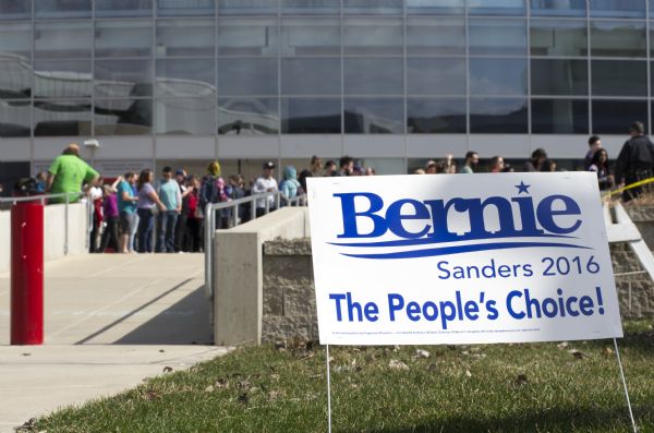 A lawn sign sitting in front of the Kohl Center reads: "Bernie Sanders 2016, The People's Choice!" In the background a line of people are waiting to enter the Kohl Center to attend the Bernie Sanders rally.