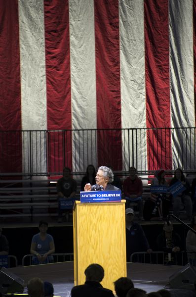 View from audience towards the mayor of Madison, Paul Soglin, standing behind a podium and speaking in the Kohl Center at the Bernie Sanders rally. There are a few people sitting on the bleachers behind him and holding signs with Sanders' campaign slogan: "A Future to Believe In." There is a large American flag hanging from the ceiling in the background.