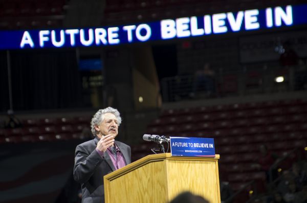 Paul Soglin, Madison's Mayor, standing behind a podium and gesturing while addressing the audience at the Bernie Sanders rally in the Kohl Center. A lighted sign high up in the background is spelling out Sanders' campaign slogan: "A Future to Believe In."