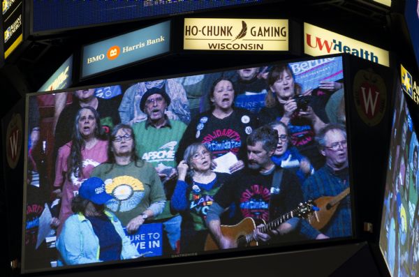 A large screen in the Kohl Center at the Bernie Sanders rally is broadcasting the Solidarity Sing Along, a progressive and pro-labor group of people who sing every weekday in the capitol building. The group is singing, and two men are playing a guitar and a mandolin. 