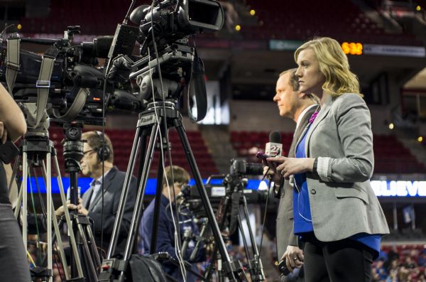 Members of the press setting up their cameras at the Bernie Sanders rally held in the Kohl Center. On the right, a woman is standing and watching, while holding her cell phone and a stylus pen. Behind her, Greg Neumann, a reporter with WKOW 27, is standing in front of a camera and holding a microphone. In the background is a lighted sign (obscured) spelling out Sanders' campaign slogan "A Future to Believe In."