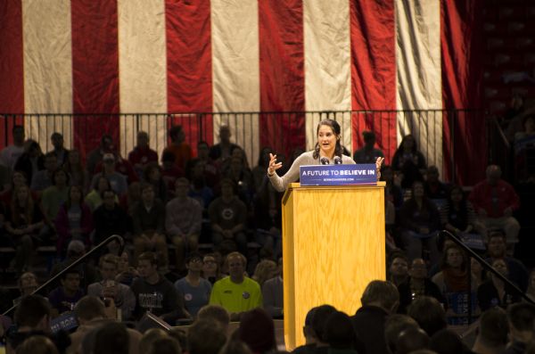 Actress Shailene Woodley standing behind a podium and addressing the crowd at the Bernie Sanders rally held in the Kohl Center.  A sign on the podium bears Sanders' campaign slogan: "A Future to Believe In."  A large American flag hangs from the ceiling in the background.