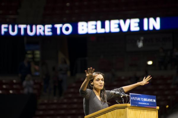 Actress Rosario Dawson standing behind a podium and gesturing while speaking at the Bernie Sanders rally held in the Kohl Center. A sign on the podium and a lighted sign high up in the background bear Sanders' campaign slogan: "A Future to Believe In."