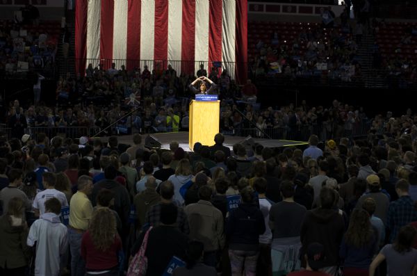 View over the large standing audience surrounding the stage at the Bernie Sanders rally held at the Kohl Center. Many people in the crowd are holding up political signs, both official and handmade. On the stage, actress Rosario Dawson is standing behind a podium and speaking to the audience. She is holding her arms above her head and forming the shape of a heart with her hands. A sign on the podium bears Sanders' campaign slogan: "A Future to Believe In." A large American flag hangs from the ceiling in the background.