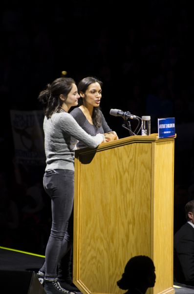 Actresses Shailene Woodley (left) and Rosario Dawson (right) standing at the podium and addressing the audience of the Bernie Sanders rally held in the Kohl Center. Two secret service agents, a man and a woman, stand below them.