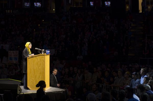 Executive Director of the Arab American Association of New York, Linda Sarsour, standing at a podium and speaking to the audience at the Bernie Sanders rally held in the Kohl Center. She is wearing a yellow and black hijab. Two secret service agents, a man and a woman, are standing in front of the podium below the stage. Behind Sarsour in the crowd a supporter is holding up a sign reading: "Fire is Catching, Bern with Us."
