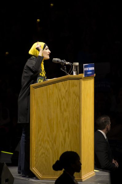 Executive Director of the Arab American Association of New York, Linda Sarsour, gesturing with her hand while speaking to the audience at the Bernie Sanders rally held in the Kohl Center. She is wearing a yellow and black hijab. Two secret service agents, a man and a woman, are standing in front of the podium below the stage.