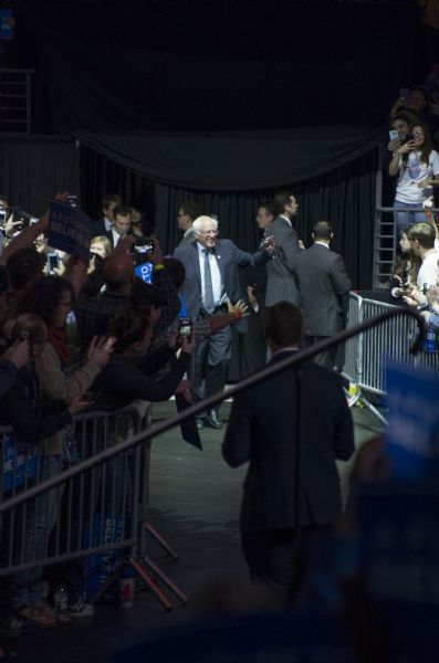 The United States Senator from Vermont, Bernie Sanders, smiling and waving while walking towards the stage at his rally held in the Kohl Center. A crowd of supporters is pressing against the guard rails as they take pictures and videos. Some are holding up signs, or waving. Secret Service agents are watching the crowd.