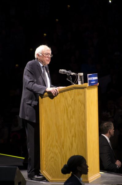 The United States Senator from Vermont, Bernie Sanders, standing at a podium and addressing the audience at his rally held in the Kohl Center. Two secret service agents, a man and a woman, stand in front of the podium below the stage.