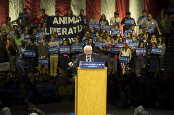 Bernie Sanders standing behind a podium and gesturing with his hand while addressing the audience at his rally held in the Kohl Center. Behind him is a crowd of supporters, mostly young men and women, who are holding signs with his campaign slogan: "A Future to Believe In." Among them, a small group is holding up a large black and white banner reading: "Animal Liberation Now." There is a large American flag in the background.