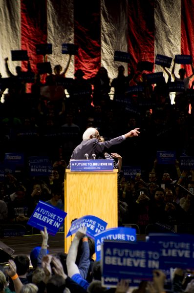 View from audience of Bernie Sanders standing behind the podium and pointing towards the right. There is someone standing with him on stage. The audience around the stage is cheering, holding up campaign signs, and taking video or pictures. A group of people in the background are silhouetted against a large American flag hanging from the ceiling.