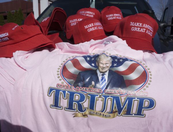 Display of Donald Trump merchandise, including several red baseball caps, red bucket hats, and a pink t-shirt, all with his campaign slogan "Make America Great Again." The shirt includes an image of Trump in front of the American flag.