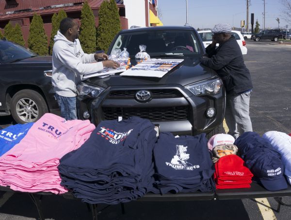 Two African American men are attaching buttons to a display on the hood of a car in a parking lot outside the Donald Trump rally. T-shirts and hats bearing Trump's image or his campaign slogan "Make America Great Again," are sitting on a long table in the foreground. One of the shirts has an illustrated image of Donald Trump holding a gun and reads: "Go Ahead Isis, Make My Day!"
