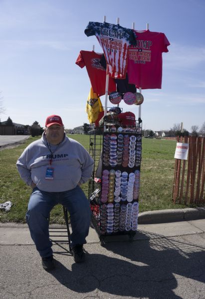 A man wearing a volunteer badge for the Janesville Donald Trump rally is sitting on a stool outdoors next to a small display of campaign merchandise. The t-shirts and hats have Trump's campaign slogan: "Make America Great Again." The buttons range from a simple image of Trump, or his name, or the Republican elephant, to anti-Hillary buttons and calls to bomb Isis. One set of buttons reads: "Trump 2016, Finally Someone with Balls."