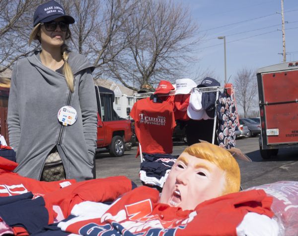 A woman is standing behind her merchandise in a parking lot outside of the Janesville Donald Trump rally. She is wearing sunglasses, and a hat bearing the Trump campaign slogan: "Make America Great Again." She is selling hats, t-shirts, and campaign buttons which are displayed on a rack behind her. The table in the foreground displays a mask of Trump's face for sale among t-shirts.
