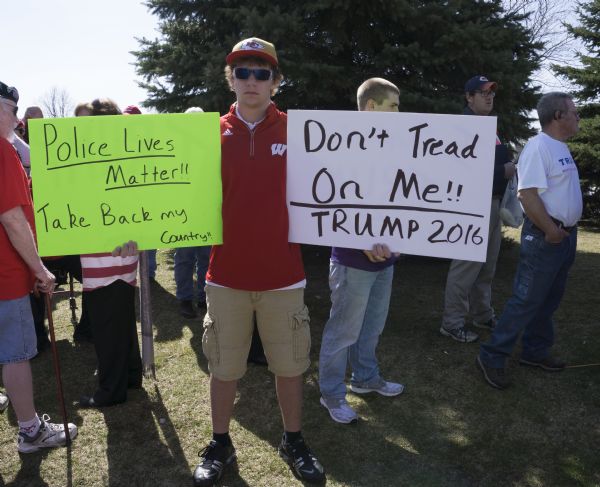 A young man is standing outdoors wearing a UW Badgers sweater, a Kansas City Chiefs cap, and sunglasses and is holding up two signs. The one on the right reads: "Don't Tread On Me!! Trump 2016." The one on the left reads: "Police Lives Matter!! Take Back my country!!" Other men are standing around him, waiting to get into the Trump rally.