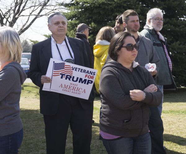 An elderly man in a suit is holding a sign reading: "Veterans for Trump, Make America Great Again!" Other men and women are standomg around him waiting to enter the Donald Trump rally.