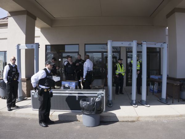 Nine Secret Service agents and police, all men, are standing around the doors at the entrance to the Janesville Conference Center which is hosting the Donald Trump rally. Three metal detectors are set up, and a large metal box is lying on the ground. Most of the men are wearing bullet proof vests.