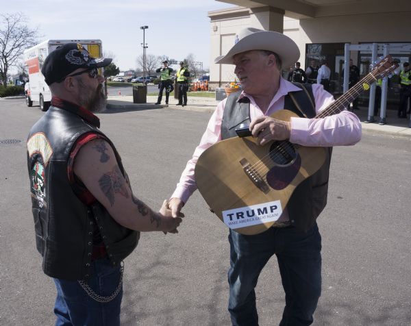 Two Donald Trump supporters are shaking hands outside of the Janesville Conference Center. The man on the right is wearing a cowboy hat and is holding a phone, and a guitar with a Trump bumper sticker on it. The other man is wearing a cap and a leather vest which has an image of a skull wearing a military helmet and reads: "Disabled Veterans." In the background police and secret service men are standing around the tables and metal detectors at the entrance.