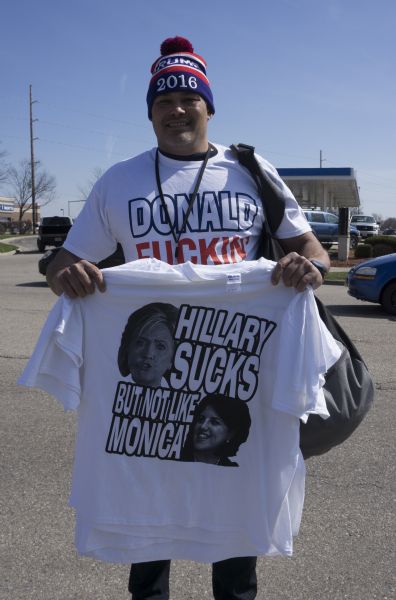 A man is posing for the camera in a parking lot holding up his merchandise while wearing a hat in red, white and blue that reads: "Trump 2016," and a white shirt that reads: "Donald Fuckin' Trump." The man is also holding up a t-shirt with images of Hillary Clinton and Monica Lewinsky that reads: "Hillary sucks but not like Monica."