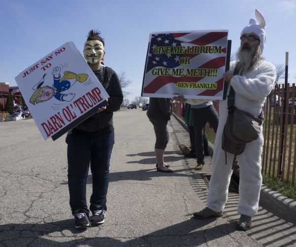 Two people are holding up their protest signs while posing for the camera outside the Donald Trump rally. The man on the right is wearing a bunny costume and is holding a sign with an image of the American flag and which reads: "Give me Librium or Give Ne Meth!!! - Ben Franklin." The other person is holding a sign with an image of Trump as Donald Duck and reads: "Just Say No To Darn Ol' Trump." This person is also wearing a Guy Fawkes mask.