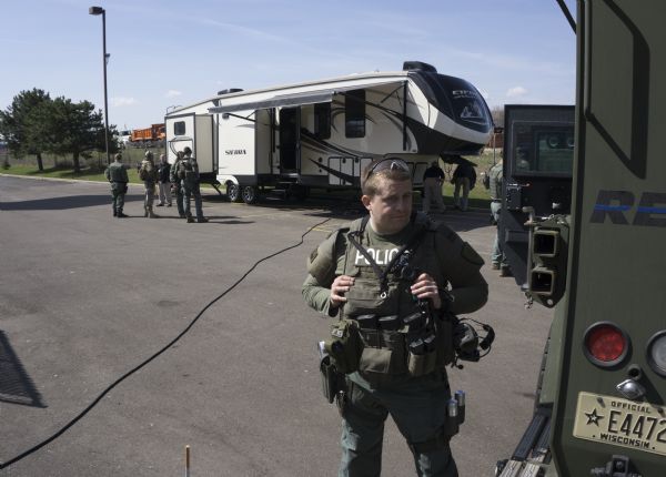A police officer in a SWAT (Special Weapons and Tactics) uniform is standing next to an armored vehicle. Other men, many also in SWAT uniforms, are standing around in the background next to armored vehicles and a large trailer.
