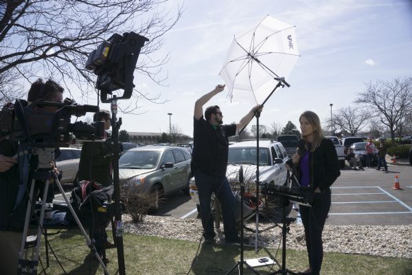NBC news correspondent Katy Tur is standing outdoors and speaking into a microphone. She is reporting on the Donald Trump rally. A camera operator is standing on the left, and a man stands in the center holding up an umbrella with the NBC logo to adjust the outdoor light. In the background on the right three men are near parked cars in the parking lot looking at Trump posters.
