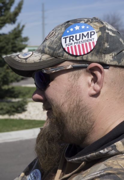 Profile of a man wearing a camouflage baseball cap, sunglasses, and sweatshirt. He has a beard and mustache. He is wearing a button on his cap that is decorated with the American flag motif and reads: "Trump for President." On his shirt, partially visible, he is wearing another button which reads "Trump bomb the hell out of..." 
