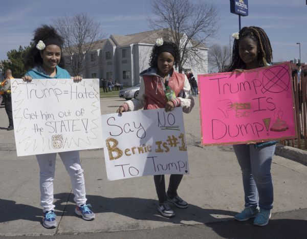 Three young African American women posing outdoors with their handmade protest signs, each one wearing a white rose in their hair. The signs read, from left to right: "Trump = Hate, Get him out of the State," "Say No to Trump, Bernie is #1," and "Trump is Dump."