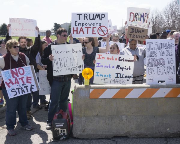 A group of protesters are holding signs while standing behind a stone barricade outside the Donald Trump rally. A few of the signs make reference to Trumps remarks on Hispanic Americans. One sign reads: "I'm not a criminal, I'm not a rapist, I'm not illegal, I matter!" while another reads: "Mexican born, Mexican tax payer!!!" One man in the crowd is holding up a pro-Trump sign reading: "Win Donld [sic] Trump, #1, Make American [sic] Great Again, that's right."