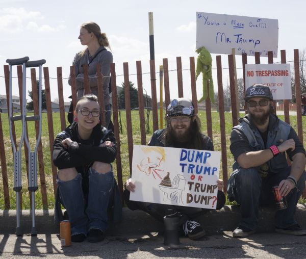 Three young people, two men and one women, are sitting on the curb in front of a fence with a sign reading: "No Trespassing By Order of Police." Behind them a woman is standing on the grass on the other side of the fence. The woman in front on the left is sitting next to crutches leaning against the fence. The man in the middle is holding a sign with an image of Trump yelling at a toilet and reading: "Dump Trump or Trump Dump?" The man on the right is sitting in front of a sign on a pole which reads: "You're a Mean One!!!! Mr. Trump," and there is a Grinch puppet attached to the fence post nearby.