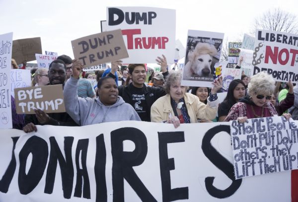 Three women and one man are holding up a large banner, only a portion of which can be read: "ionaires." They are also holding their own signs, as do many in the crowd of protesters behind them. Many of the signs read: "Dump Trump." The two women on the right are wearing white roses on their clothing. 