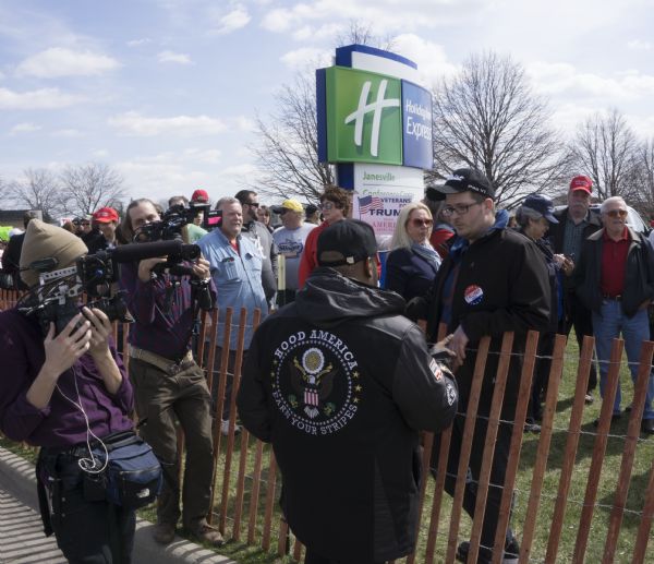 Standing in the foreground in front of a fence are two people with cameras filming an African American man talking to a group of people who are Trump supporters standing on the other side of the fence waiting to enter the Janesville Conference Center for the rally. The man is wearing a jacket, and on the back is printed: "Hood America, Ear Your Stripes."